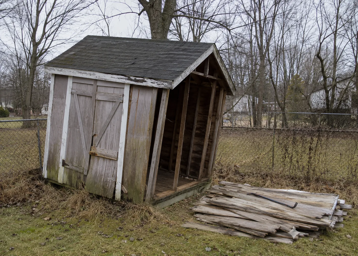 Old leaning wooden backyard shed with boards stacked on the grass beside it